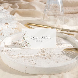 May include: A rectangular white place card featuring a delicate floral illustration on the left side and the black printed text Lucas Anderson TABLE 01. The card is positioned upright on a round white tray with gold-toned marble veining.