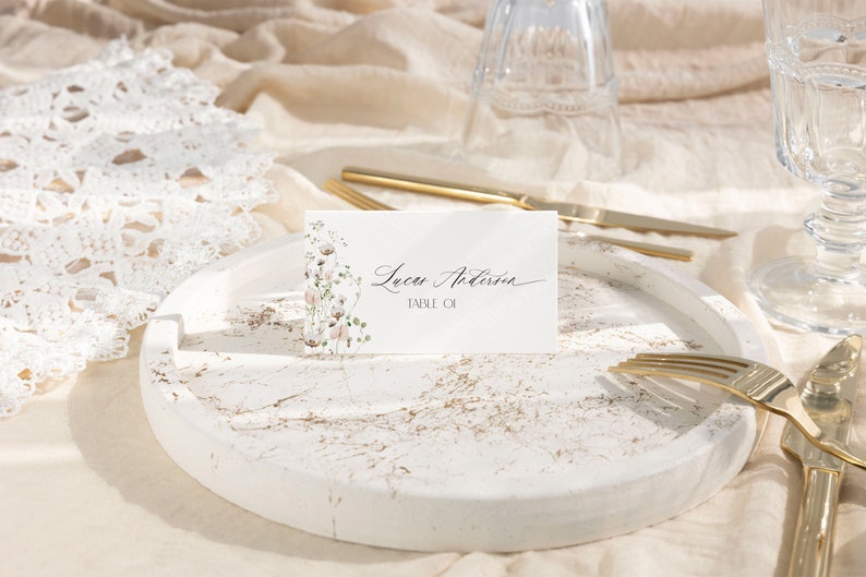 May include: A rectangular white place card featuring a delicate floral illustration on the left side and the black printed text Lucas Anderson TABLE 01. The card is positioned upright on a round white tray with gold-toned marble veining.