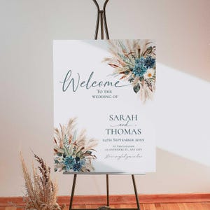 May include: A white wedding welcome sign on a black easel. The sign features the word "Welcome" in cursive, with floral arrangements in the corners. The names "Sarah and Thomas" and the wedding date are also displayed.