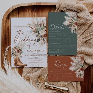 May include: A wedding invitation suite featuring a white invitation card with a brown cowboy boot and floral arrangement, a dark green details card, and a rust-colored RSVP card. All cards feature dried floral accents and elegant script text.