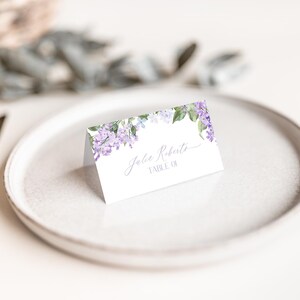May include: A white place card with a floral design featuring purple flowers and green leaves. The text "John Roberts, Table 01" is printed on the card.
