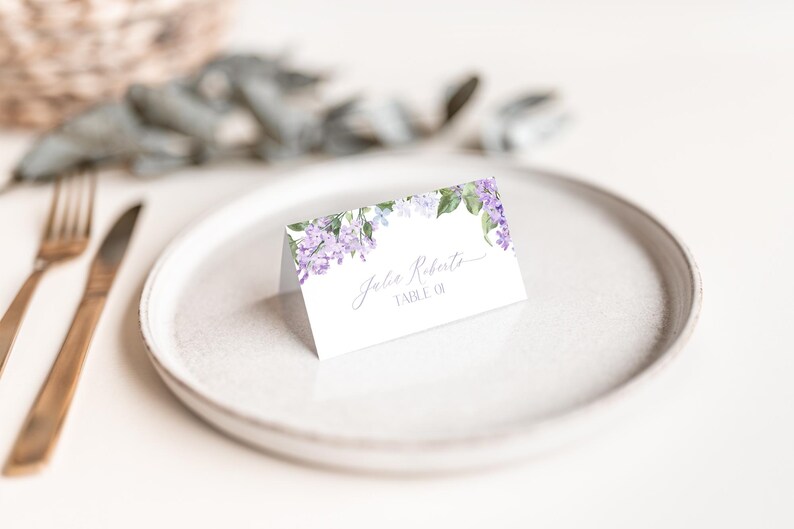 May include: A white place card with a floral design featuring purple flowers and green leaves. The text "John Roberts, Table 01" is printed on the card.