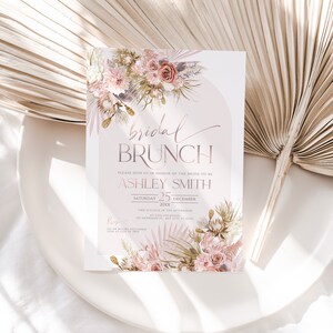 May include: A blush pink and gold bridal brunch invitation with a floral design. The invitation reads "bridal brunch" and includes the bride-to-be's name, Ashley Smith, the date, Saturday, December 25, 20xx, and the time, two o'clock in the afternoon. The invitation also includes the location, 123 Anywhere St, Any City, 31 12148. The RSVP information is on the bottom of the invitation.