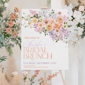 May include: A white sign with a floral design in shades of pink, purple, and yellow. The sign reads "Welcome to Sarah's Bridal Brunch" and includes the date "Saturday, December 24th" and a location. The sign is displayed on a wooden easel.