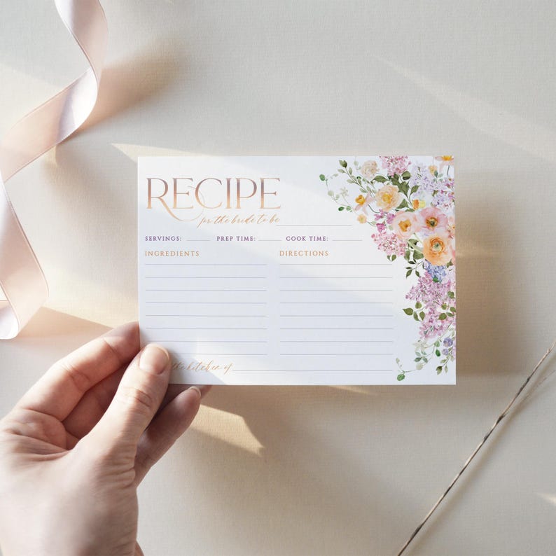 May include: A white recipe card with a floral design in shades of pink, purple, and yellow. The card has the words "RECIPE for the bride to be" printed in gold lettering. The card has sections for servings, prep time, cook time, ingredients, and directions.