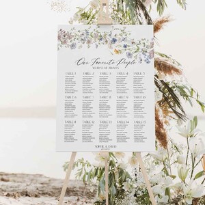 May include: A white wedding seating chart with a floral design and the text "Our Favorite People Your Seat Awaits" at the top. The chart lists tables 1 through 15 with names of guests assigned to each table. The chart is displayed on a wooden easel.