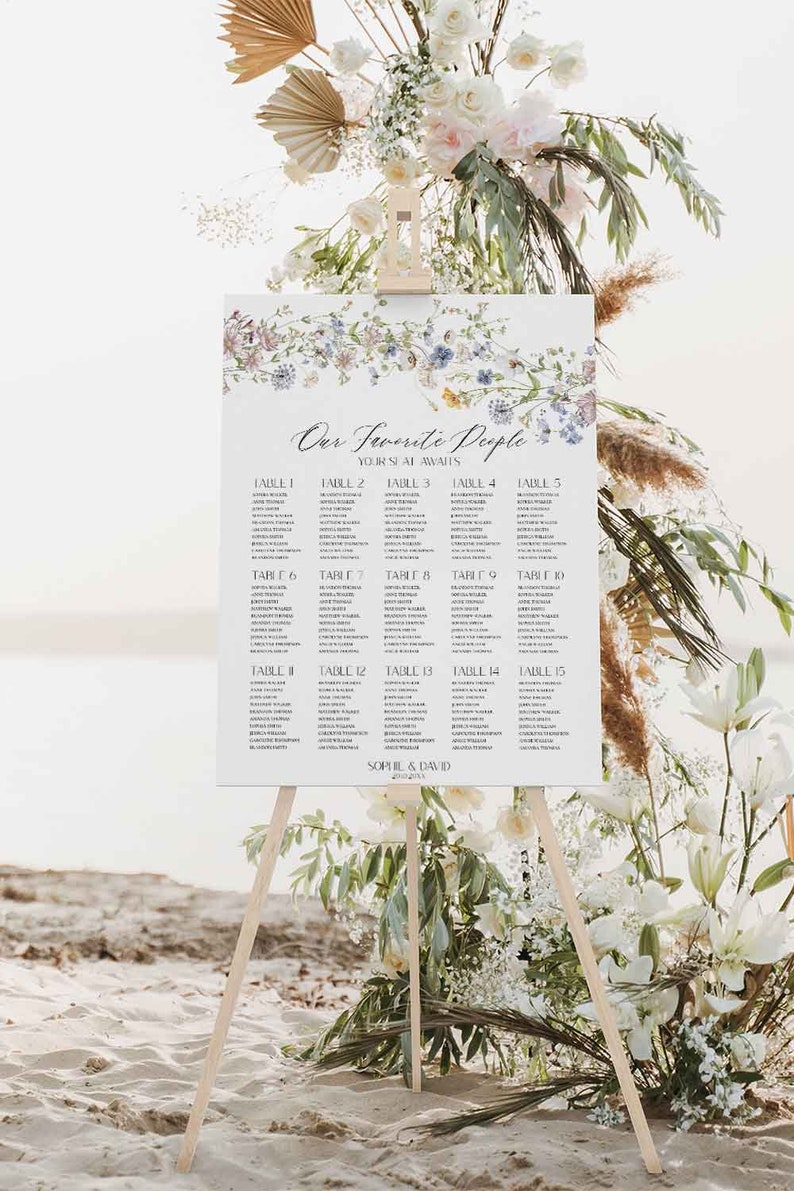 May include: A white wedding seating chart with a floral design and the text "Our Favorite People Your Seat Awaits" at the top. The chart lists tables 1 through 15 with names of guests assigned to each table. The chart is displayed on a wooden easel.