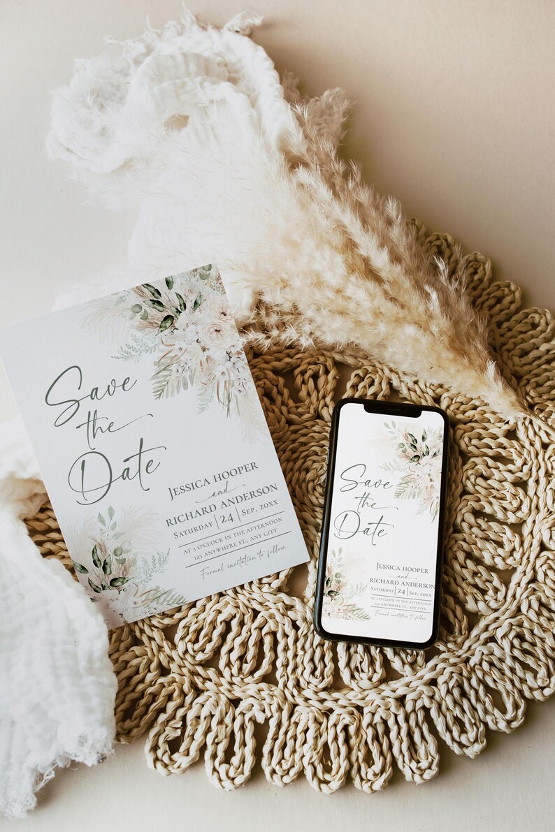 May include: A save the date card with a watercolor floral design in shades of green and white. The card reads "Save the Date" with the names Jessica Hooper and Richard Anderson, the date September 24, 20XX, and the time 1 o'clock in the afternoon. The card is sitting on a woven placemat with a bouquet of dried pampas grass.
