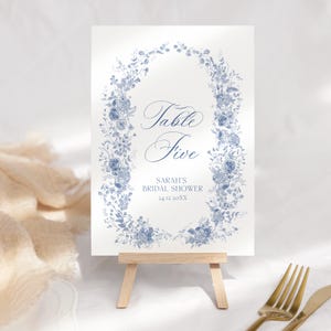 May include: A white table sign with blue floral design, displayed on a small wooden easel. The sign reads "Table Five" and "Sarah's Bridal Shower 24.12.20XX". The floral design forms an oval around the text.