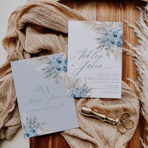 K&ouml;nnte beinhalten: Zwei Hochzeitseinladungen mit blauen und wei&szlig;en Blumenmustern. Die Einladungen liegen auf einer Holzoberfl&auml;che mit einem beigefarbenen Stoff&uuml;berzug. Der Text auf den Einladungen lautet "Ashley + Jason" und "Ashley + Jason" mit dem Datum "Sonntag, 24. Juli 20XX".