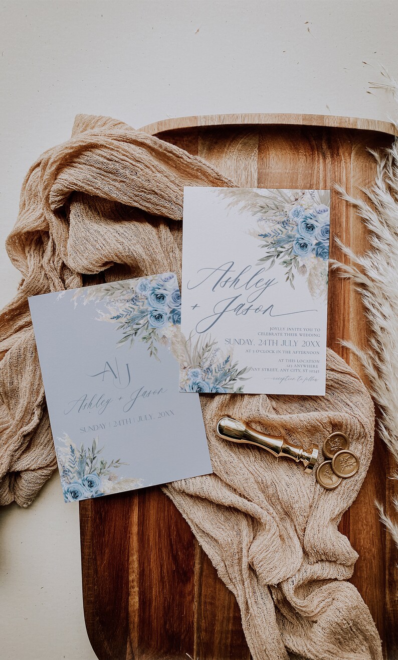 K&ouml;nnte beinhalten: Zwei Hochzeitseinladungen mit blauen und wei&szlig;en Blumenmustern. Die Einladungen liegen auf einer Holzoberfl&auml;che mit einem beigefarbenen Stoff&uuml;berzug. Der Text auf den Einladungen lautet "Ashley + Jason" und "Ashley + Jason" mit dem Datum "Sonntag, 24. Juli 20XX".