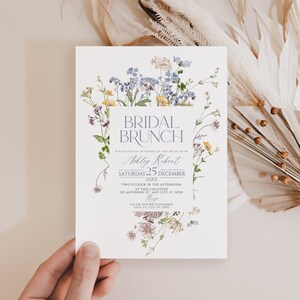 May include: A floral bridal brunch invitation with a watercolor design of blue and yellow flowers. The invitation reads "Bridal Brunch" in a blue script font and includes the bride-to-be's name, the date, and the time of the event. RSVP information is also included.