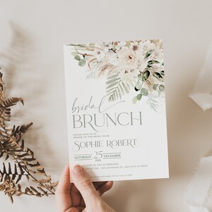 Może przedstawiać: Biało-zielone kwiatowe zaproszenie na brunch ślubny z tekstem "bridal brunch" w szarym, ozdobnym piśmie. Zaproszenie zawiera imię przyszłej panny młodej, "Sophie Robert", datę "sobota 25 grudnia 20XX" i godzinę "O'clock in the afternoon".