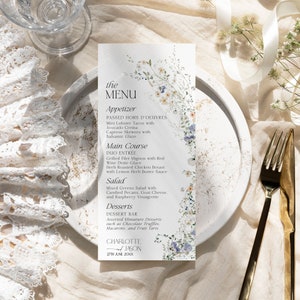 Puede incluir: Una tarjeta de men&uacute; de boda con un dise&ntilde;o floral. El men&uacute; incluye aperitivos, plato principal, ensalada y postres. La tarjeta est&aacute; decorada con delicadas flores azules y blancas y vegetaci&oacute;n. El texto en la tarjeta dice "the MENU", "Appetizer", "Main Course", "Salad", "Desserts", "CHARLOTTE and JASON", y "27TH JUNE 20XX".