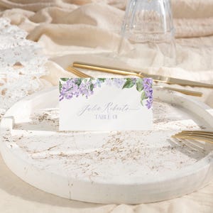 May include: A white place card with a watercolor design of purple flowers and green leaves. The text on the card reads "Julie Roberts, Table 01."
