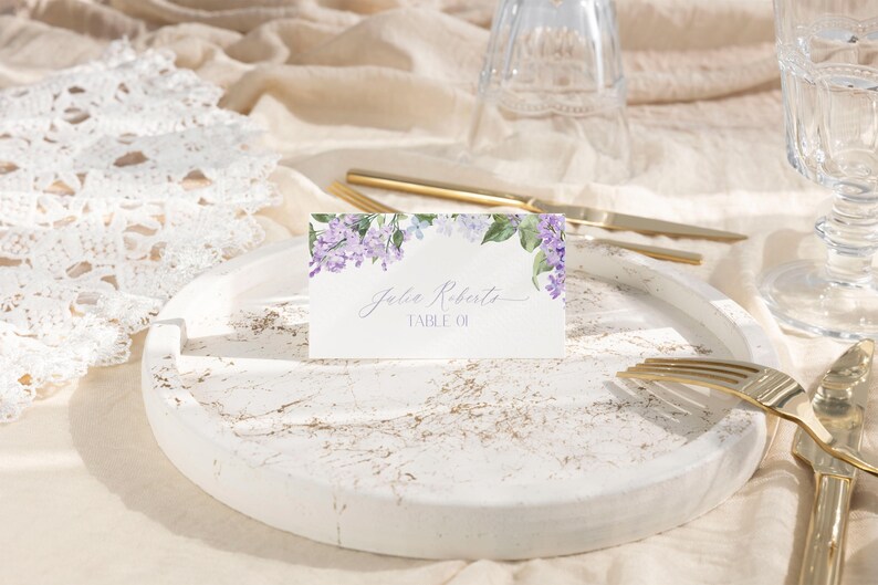 May include: A white place card with a watercolor design of purple flowers and green leaves. The text on the card reads "Julie Roberts, Table 01."