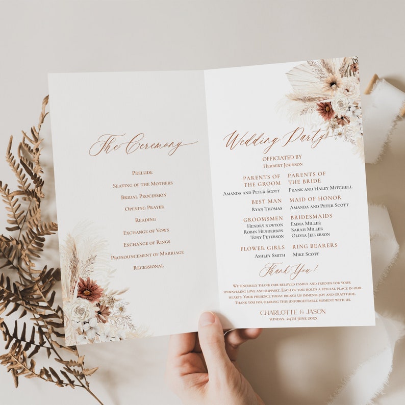 May include: A wedding ceremony programme with a floral design featuring dried pampas grass and blush-coloured flowers. The programme lists the order of events for the ceremony and the wedding party members. The text reads "The Ceremony" and "Wedding Party".