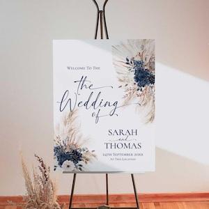 Könnte beinhalten: Ein weißes Hochzeits-Willkommensschild auf einer Staffelei. Das Schild zeigt eine elegante Kalligraphie mit dem Text "Willkommen zur Hochzeit von Sarah und Thomas", dem Datum und dem Ort. Marineblaue und beige Blumendesigns schmücken die Ecken.