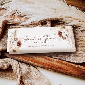 May include: A white chocolate bar with a floral design and the text "Sarah & Thomas" and "09.24.20XX" in brown lettering. The text "Thank you for celebrating with us!" is also included in brown lettering.
