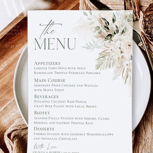 May include: A white rectangular menu card featuring dark green text and a floral illustration of pampas grass and white roses. The text lists appetizers, main course, beverages, buffet, and desserts, ending with Olivia & Robin 10.11.20XX.