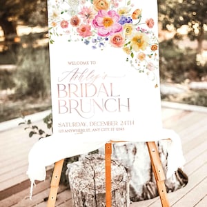 May include: A white rectangular sign on a wooden easel featuring a watercolor floral arrangement in pink, orange, and yellow. The text reads: WELCOME TO Ashley's BRIDAL BRUNCH SATURDAY, DECEMBER 24TH 123 ANYWHERE ST., ANY CITY, ST 12345.