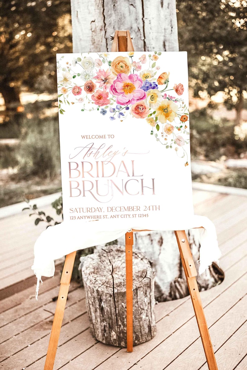 May include: A white rectangular sign on a wooden easel featuring a watercolor floral arrangement in pink, orange, and yellow. The text reads: WELCOME TO Ashley's BRIDAL BRUNCH SATURDAY, DECEMBER 24TH 123 ANYWHERE ST., ANY CITY, ST 12345.