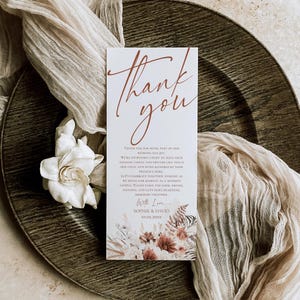 May include: A rectangular wedding thank you card with white background and rust-colored script text reading Thank you. The card features printed floral illustrations at the bottom and text including the names Sophie and David with date 10.02.20XX.