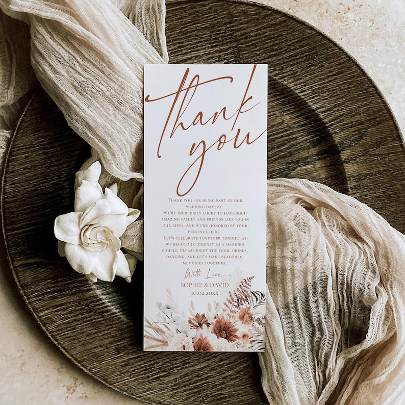 May include: A rectangular wedding thank you card with white background and rust-colored script text reading Thank you. The card features printed floral illustrations at the bottom and text including the names Sophie and David with date 10.02.20XX.