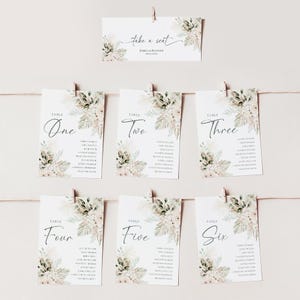 Puede incluir: Seis tarjetas de mesa con dise&ntilde;os florales y los nombres de los invitados listados en cada tarjeta. Las tarjetas est&aacute;n colgadas de una cuerda con pinzas para la ropa. La tarjeta superior dice "Take a Seat" con los nombres Jessica & Richard debajo.