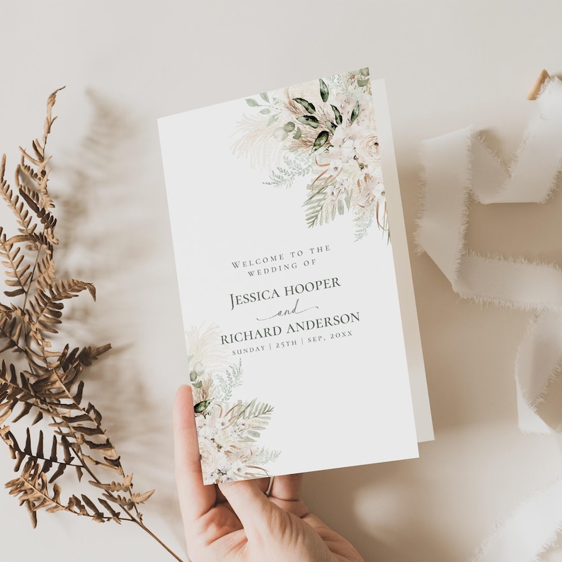 May include: A white wedding program card featuring a floral and pampas grass illustration in the top right and bottom left corners. The text reads: Welcome to the wedding of Jessica Hooper and Richard Anderson, Sunday 25th Sep, 20xx.