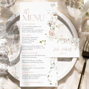 May include: A white menu card with floral illustrations and pink text listing appetizers, salads, main courses, beverages, and desserts for Sarah's Bridal Shower. A smaller matching place card with the name Julia Roberts is placed on top.