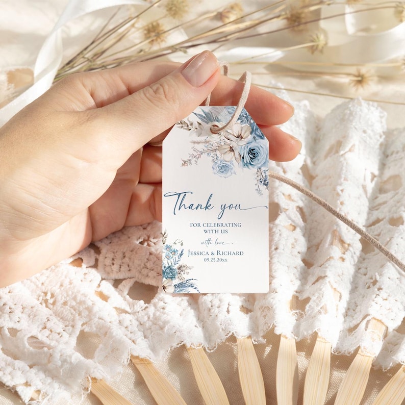 Pu&ograve; includere: Un'etichetta rettangolare in carta bianca con un foro nella parte superiore e un cordino beige. L'etichetta presenta illustrazioni floreali blu e beige e il testo: Thank you FOR CELEBRATING WITH US with love JESSICA & RICHARD 09.25.20xx.