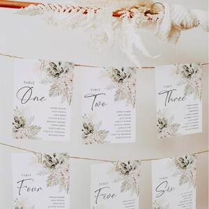 Puede incluir: Seis tarjetas blancas con dise&ntilde;os florales y clips dorados colgando de una cuerda. Cada tarjeta tiene un n&uacute;mero de mesa, del uno al seis, y una lista de nombres. Las tarjetas est&aacute;n contra una pared blanca.