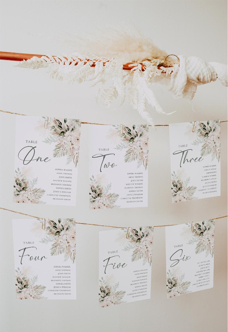 Puede incluir: Seis tarjetas blancas con dise&ntilde;os florales y clips dorados colgando de una cuerda. Cada tarjeta tiene un n&uacute;mero de mesa, del uno al seis, y una lista de nombres. Las tarjetas est&aacute;n contra una pared blanca.