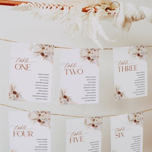 Puede incluir: Seis tarjetas de mesa blancas con nombres, sujetas con clips a una cuerda, se muestran sobre un fondo blanco. Cada tarjeta tiene un dise&ntilde;o floral y la palabra "Mesa" seguida de un n&uacute;mero en marr&oacute;n. Flores secas y hierba de la pampa est&aacute;n dispuestas encima.