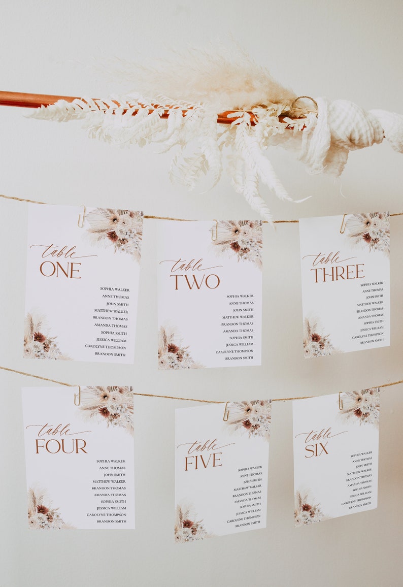 Puede incluir: Seis tarjetas de mesa blancas con nombres, sujetas con clips a una cuerda, se muestran sobre un fondo blanco. Cada tarjeta tiene un dise&ntilde;o floral y la palabra "Mesa" seguida de un n&uacute;mero en marr&oacute;n. Flores secas y hierba de la pampa est&aacute;n dispuestas encima.