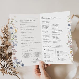 May include: A white wedding program card featuring floral illustrations in blue, purple, and yellow along the sides. The text includes sections for The Ceremony, The Wedding Party, and a Wedding Timeline, ending with a Thank You note from Sophie and David.