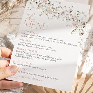May include: A white rectangular menu card featuring a delicate floral illustration at the top and rose gold text. The menu lists appetizers, salads, main courses, beverages, and desserts, concluding with the text Sarah's Bridal Shower 4.12.20XX.
