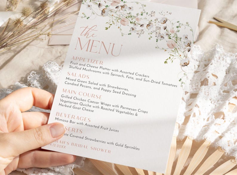 May include: A white rectangular menu card featuring a delicate floral illustration at the top and rose gold text. The menu lists appetizers, salads, main courses, beverages, and desserts, concluding with the text Sarah's Bridal Shower 4.12.20XX.
