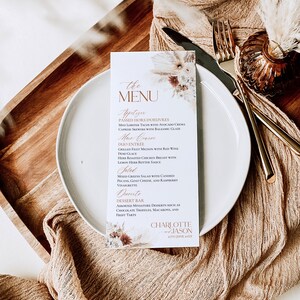 May include: A rectangular white paper menu card featuring floral illustrations and the text THE MENU, Appetizer, Main Course, Salad, Desserts, and Charlotte and Jason 27th June 20XX, placed on a round white plate with gold cutlery.