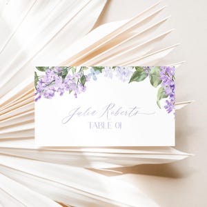 May include: Lilac floral wedding table card with the name "Julia Roberts" and the text "Table 01" in a cursive font.