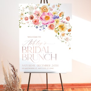 May include: A white rectangular sign on a black metal easel featuring a watercolor floral arrangement in pink, yellow, and orange at the top. The text reads: WELCOME TO Ashley's BRIDAL BRUNCH SATURDAY, DECEMBER 24TH 123 ANYWHERE ST., ANY CITY, ST 12345.