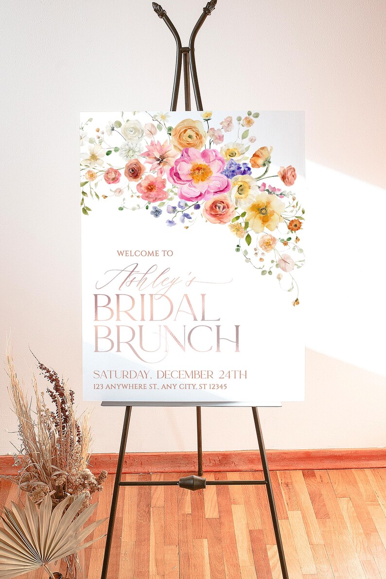 May include: A white rectangular sign on a black metal easel featuring a watercolor floral arrangement in pink, yellow, and orange at the top. The text reads: WELCOME TO Ashley's BRIDAL BRUNCH SATURDAY, DECEMBER 24TH 123 ANYWHERE ST., ANY CITY, ST 12345.