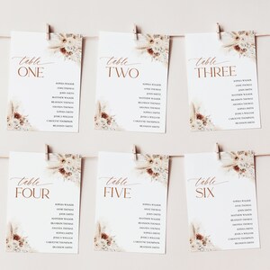 Puede incluir: Seis tarjetas de mesa blancas con nombres, sujetas a una cuerda. Cada tarjeta tiene un dise&ntilde;o floral en la esquina superior derecha y la palabra "mesa" seguida de un n&uacute;mero en color cobre. Las tarjetas son para una boda o evento.