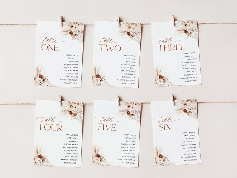 Puede incluir: Seis tarjetas de mesa blancas con nombres, sujetas a una cuerda. Cada tarjeta tiene un dise&ntilde;o floral en la esquina superior derecha y la palabra "mesa" seguida de un n&uacute;mero en color cobre. Las tarjetas son para una boda o evento.