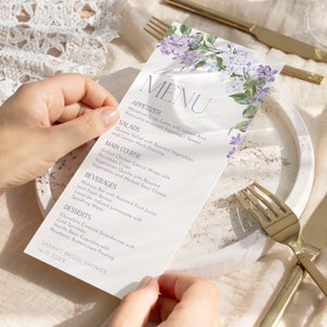 Op de afbeelding: Een wit menu met een bloemenontwerp met paarse bloemen. Het menu bevat voorgerechten, salades, hoofdgerecht, dranken en desserts. De tekst op het menu luidt "MENU", "APPETIZER", "SALADS", "MAIN COURSE", "BEVERAGES", "DESSERTS", "SARAH'S BRIDAL SHOWER" en "14.12.20XX".