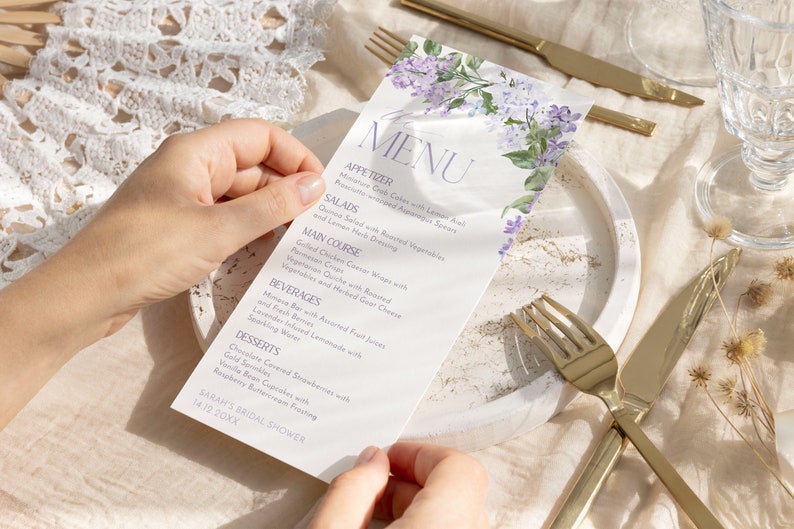 Op de afbeelding: Een wit menu met een bloemenontwerp met paarse bloemen. Het menu bevat voorgerechten, salades, hoofdgerecht, dranken en desserts. De tekst op het menu luidt "MENU", "APPETIZER", "SALADS", "MAIN COURSE", "BEVERAGES", "DESSERTS", "SARAH'S BRIDAL SHOWER" en "14.12.20XX".