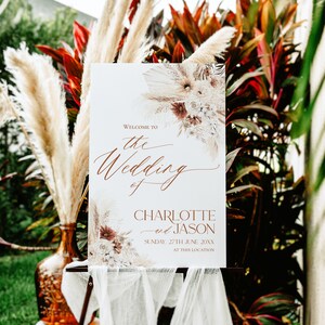May include: A white rectangular wedding welcome sign featuring copper-colored text that reads Welcome to the Wedding of Charlotte and Jason Sunday, 27th June 20XX at this location, decorated with dried floral arrangements.