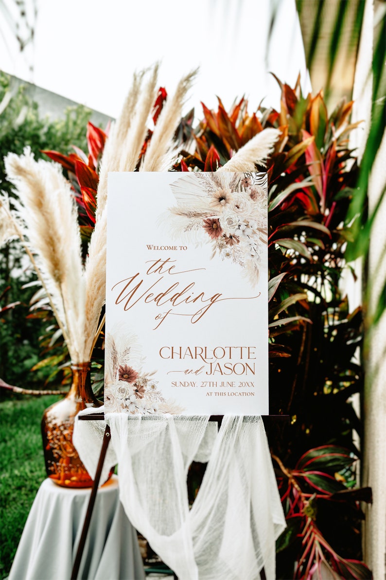 May include: A white rectangular wedding welcome sign featuring copper-colored text that reads Welcome to the Wedding of Charlotte and Jason Sunday, 27th June 20XX at this location, decorated with dried floral arrangements.