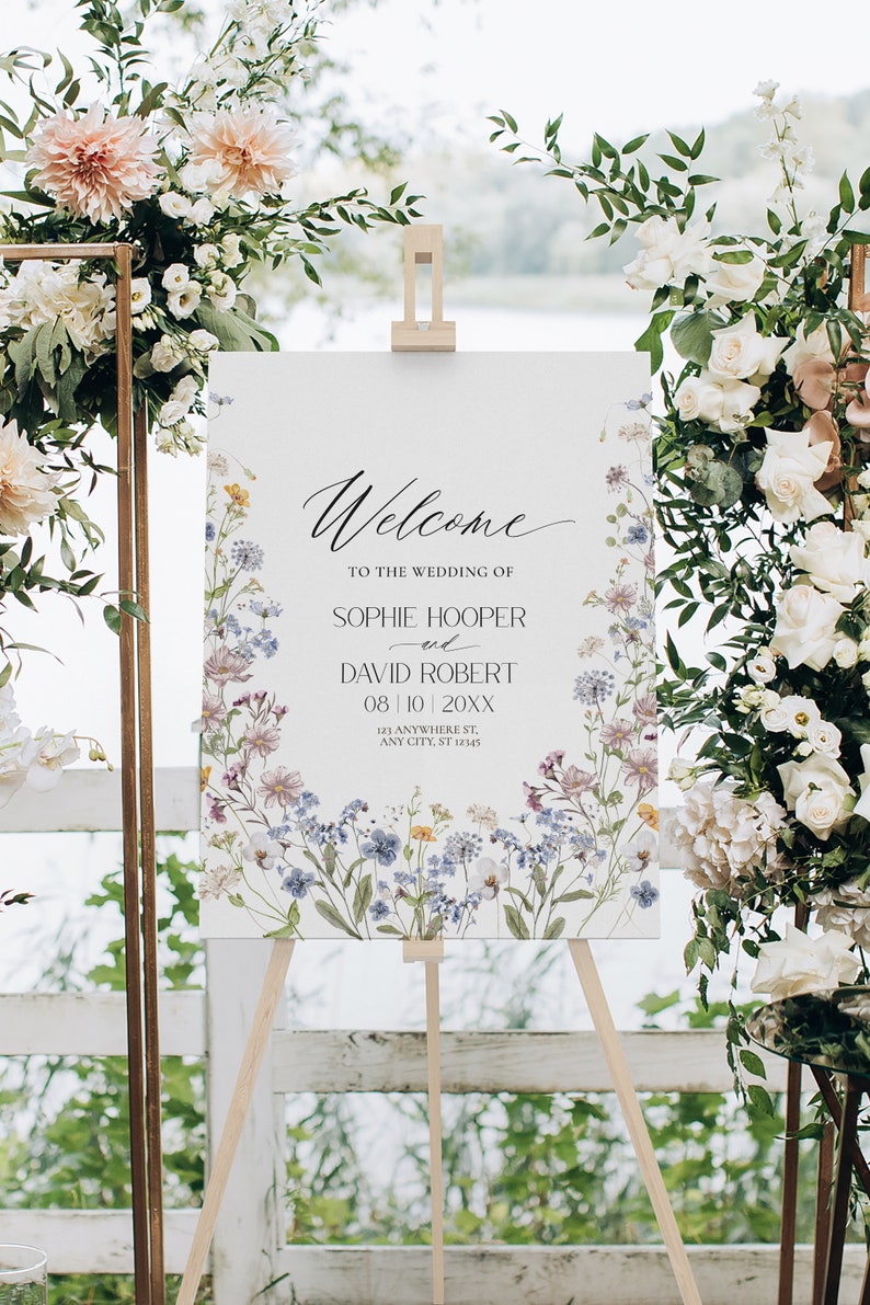 May include: A white wedding welcome sign with a floral design featuring blue, pink, and yellow flowers. The sign reads "Welcome to the wedding of Sophie Hooper and David Robert 08/10/20XX 123 Anywhere St, Any City, ST 12345".